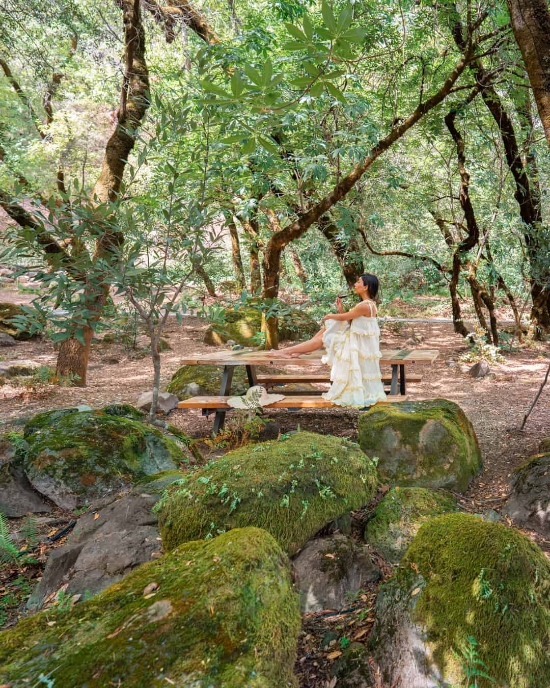 Woman in white dress sitting on picnic bench in forest.