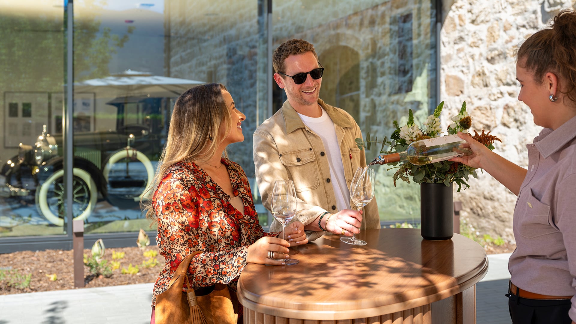 Server pours wine for smiling couple at outdoor table.