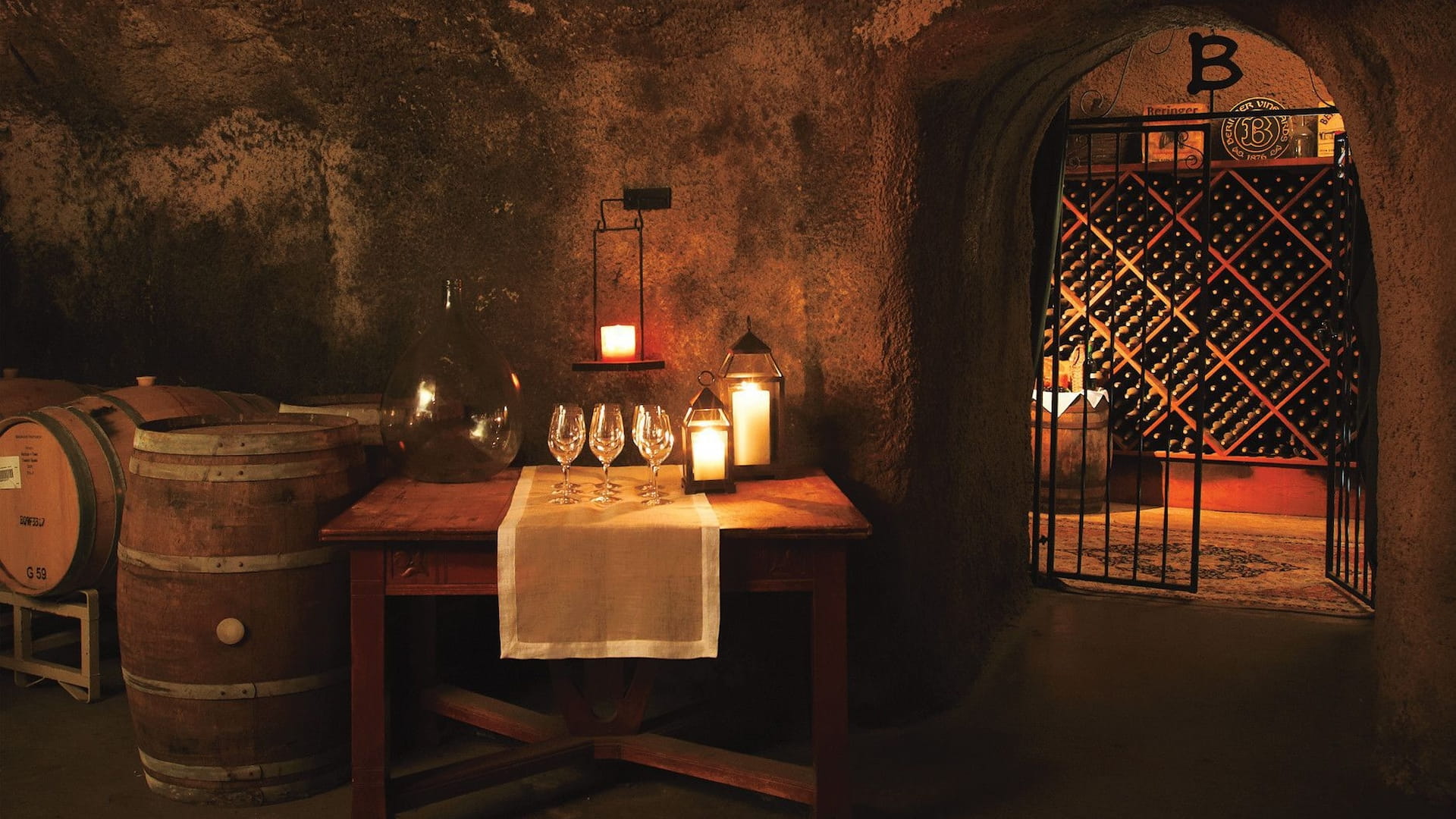 Candlelit wine cellar with wooden barrels and table.