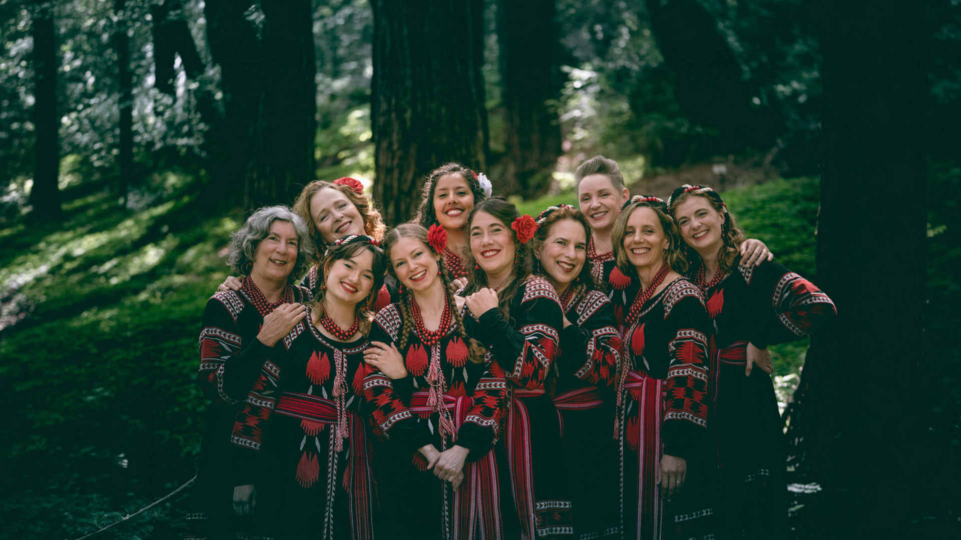 Women in matching embroidered dresses pose in a forest.