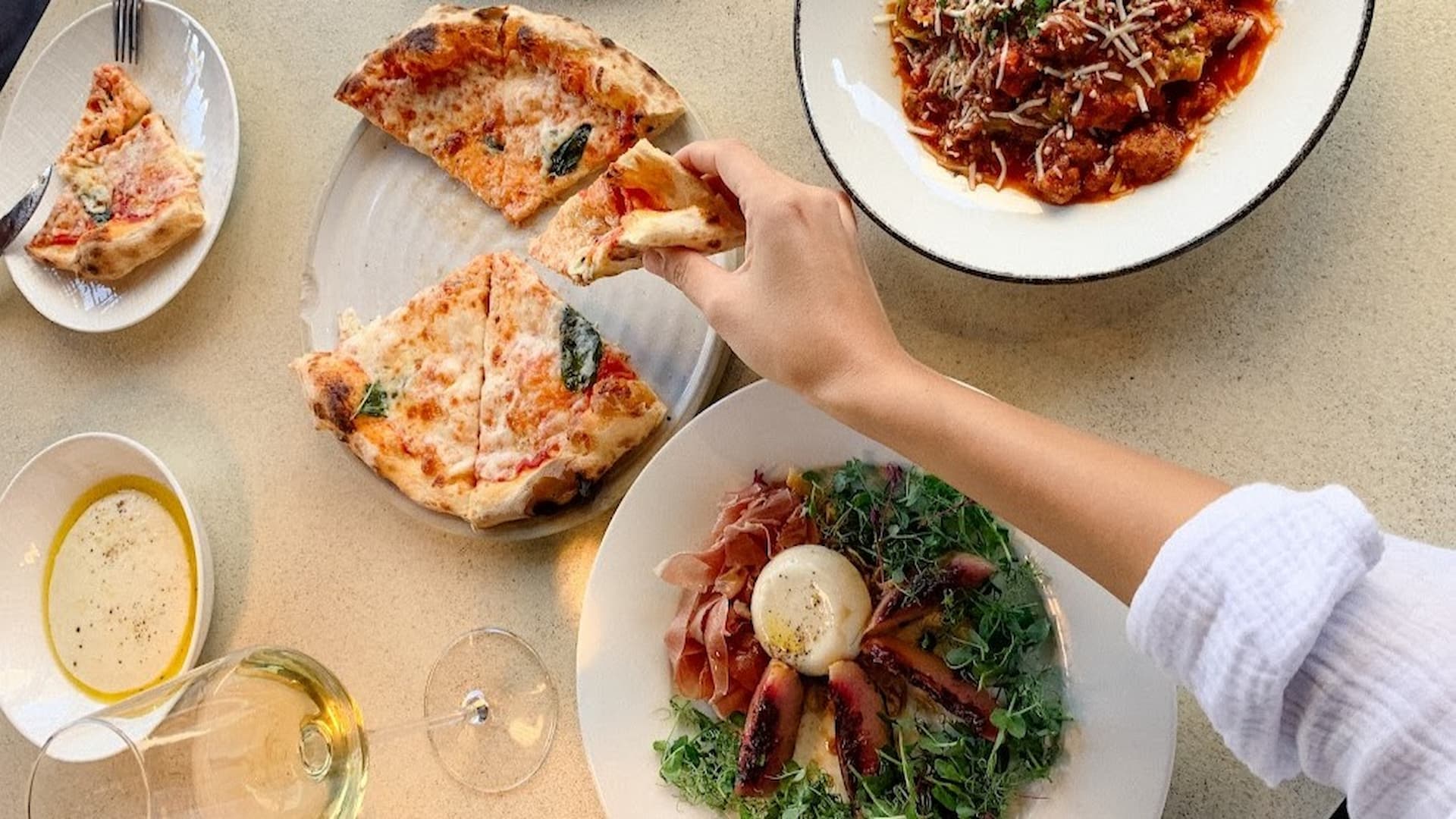 Hand reaching for pizza, pasta, and salad on table.