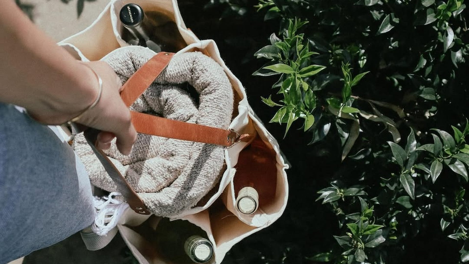 Person carrying bag with wine and blanket outdoors.