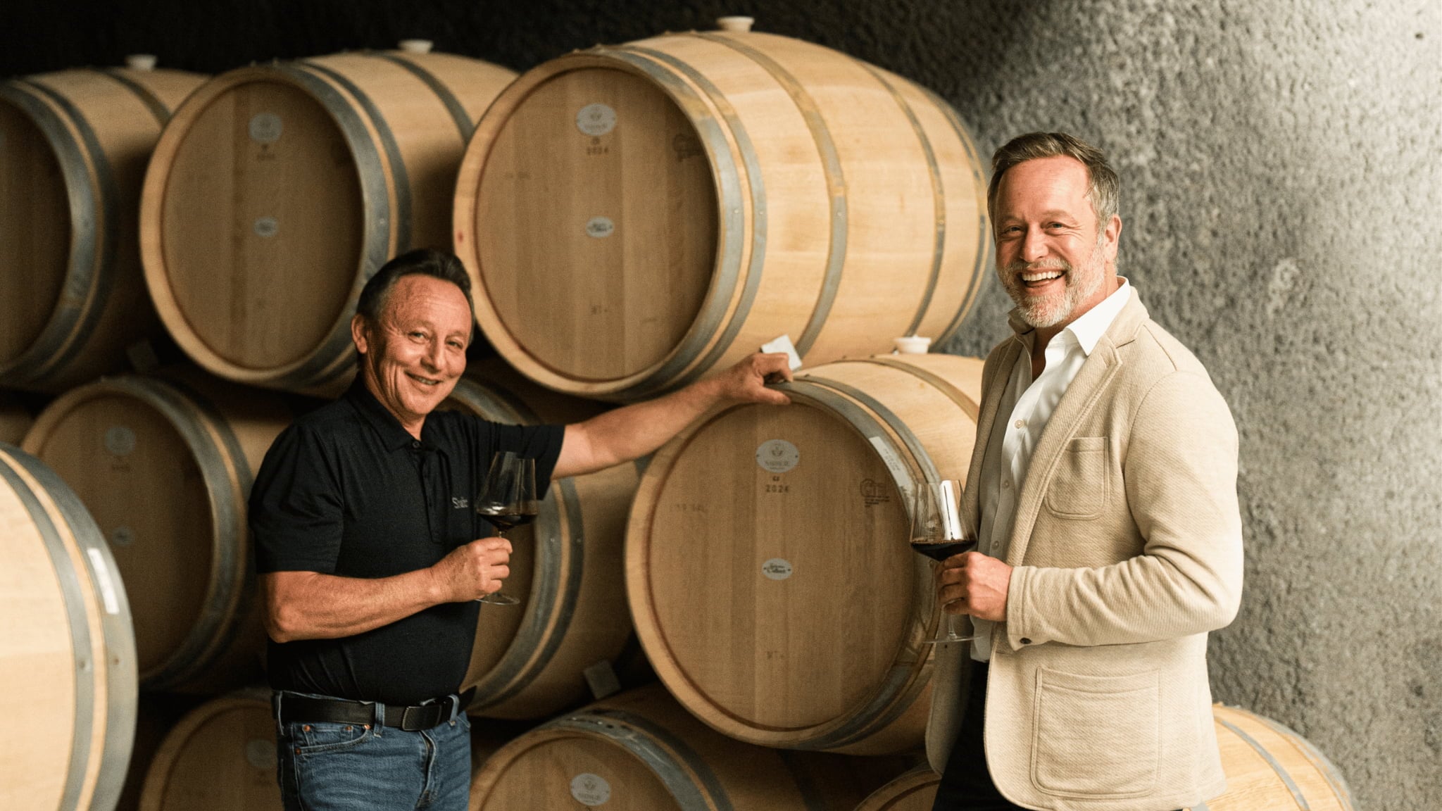 Two men tasting wine by oak barrels
