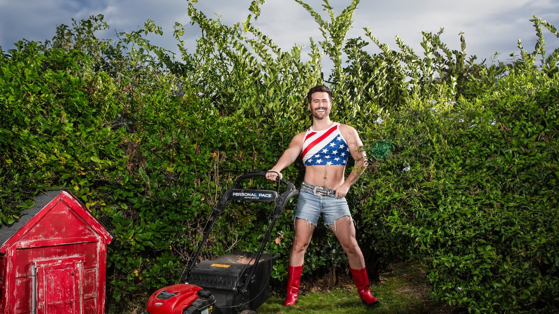 Man with lawnmower in patriotic outfit, smiling outdoors.