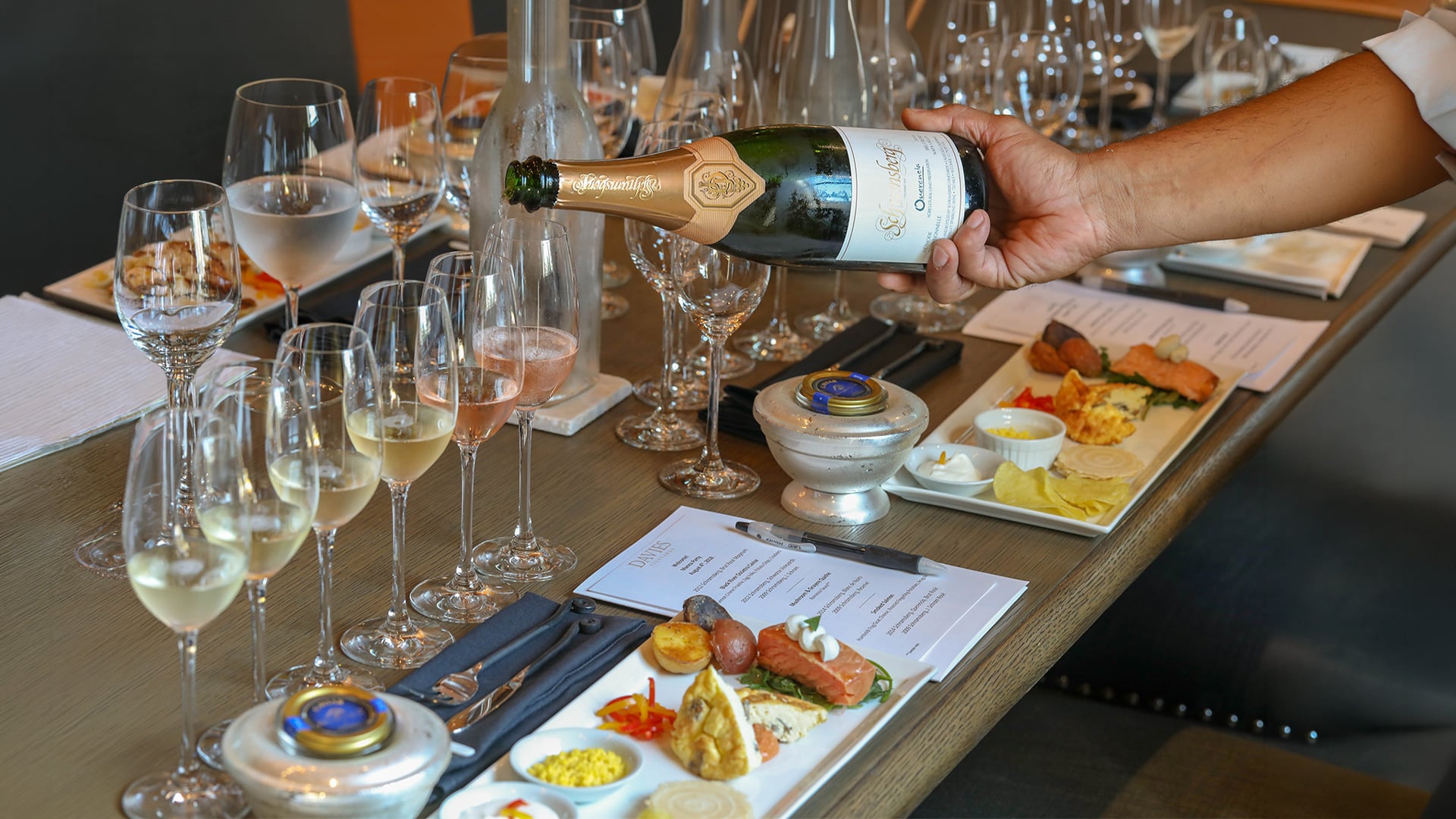Server pours sparkling wine beside tasting glasses and appetizers.