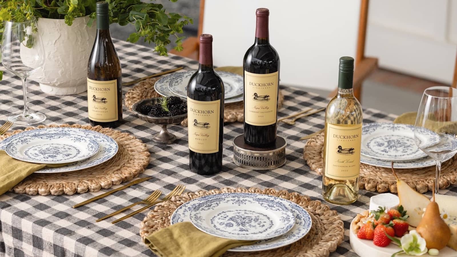 Wine bottles on checkered table with plates and fruit.