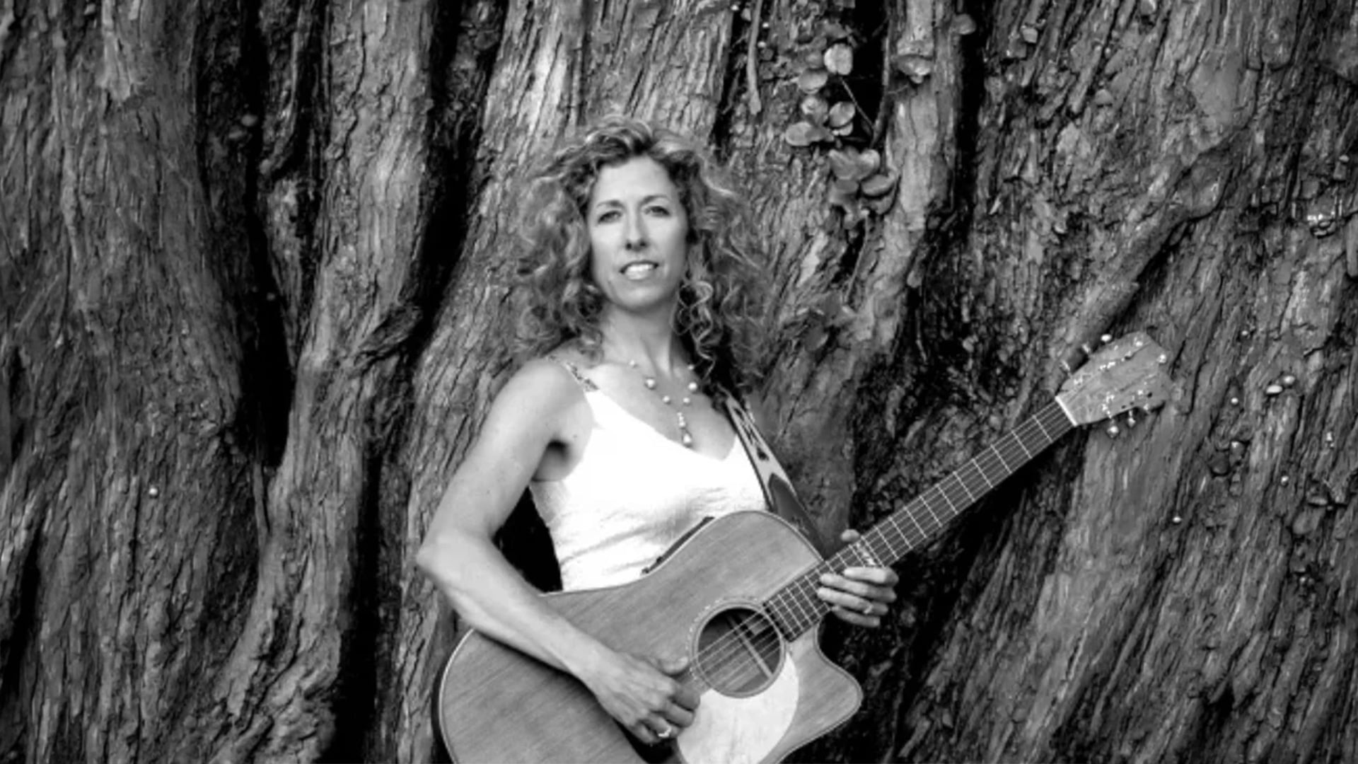 Woman holding acoustic guitar beside massive tree trunk