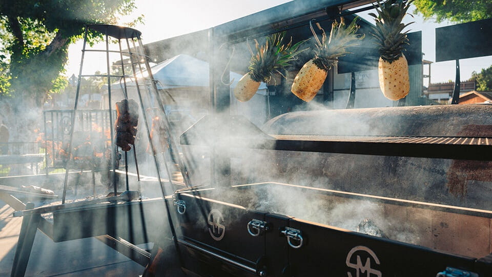 Outdoor barbecue with hanging pineapples and grilling meat.