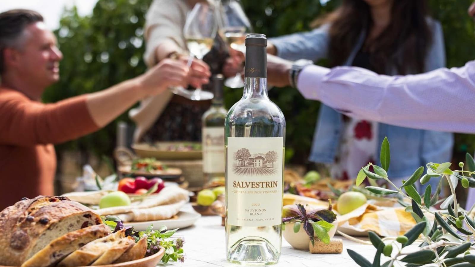 Friends toasting wine outdoors with fresh bread and greenery.