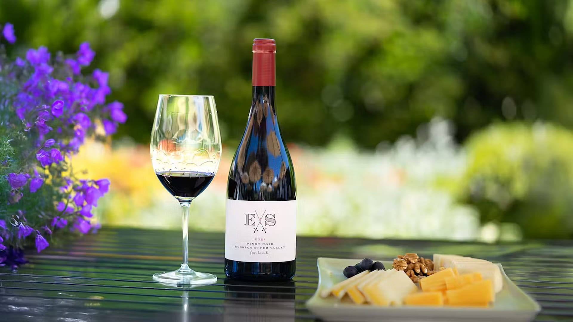 Wine, glass, cheese, and flowers on outdoor table.