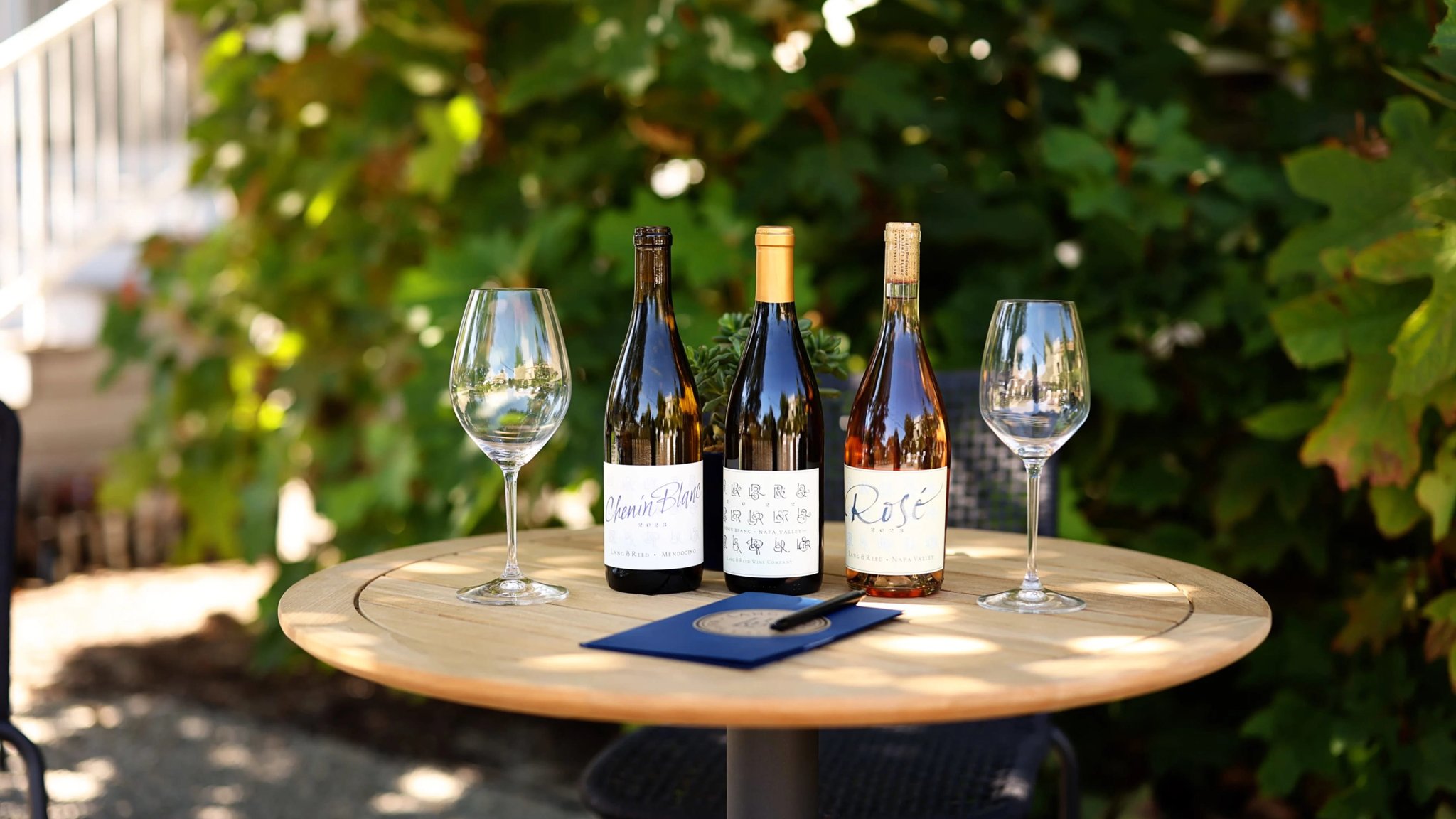Three wine bottles on outdoor table with glasses.
