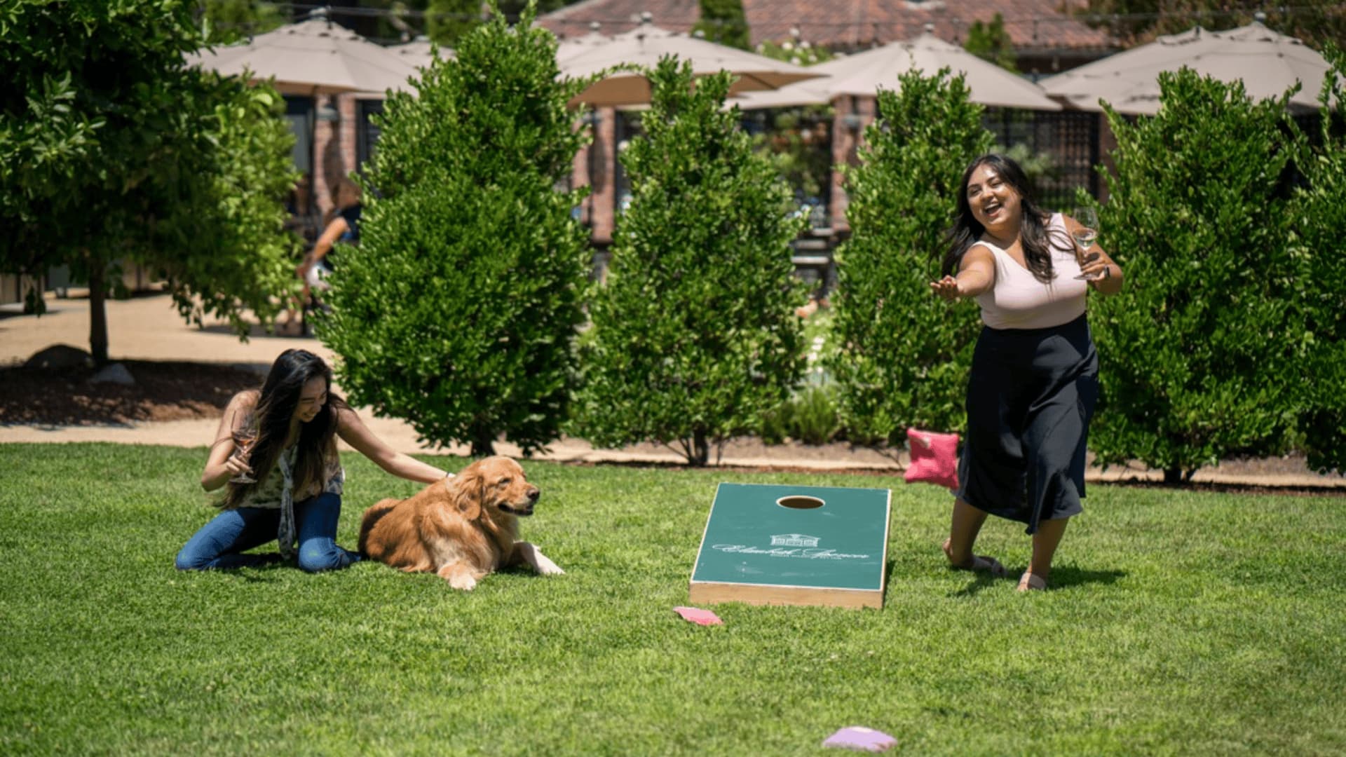 Women playing cornhole game on sunny lawn.