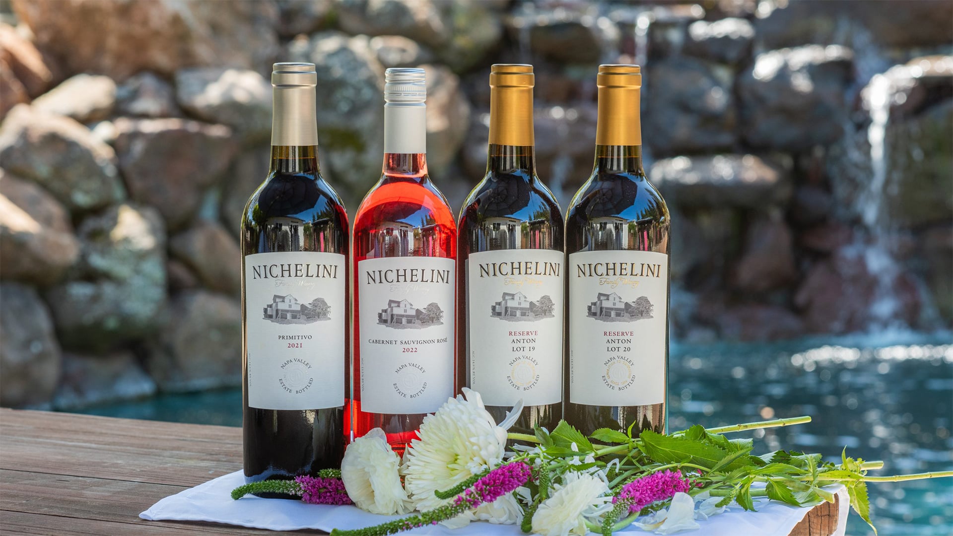 Four Nichelini wine bottles beside flowers and waterfall