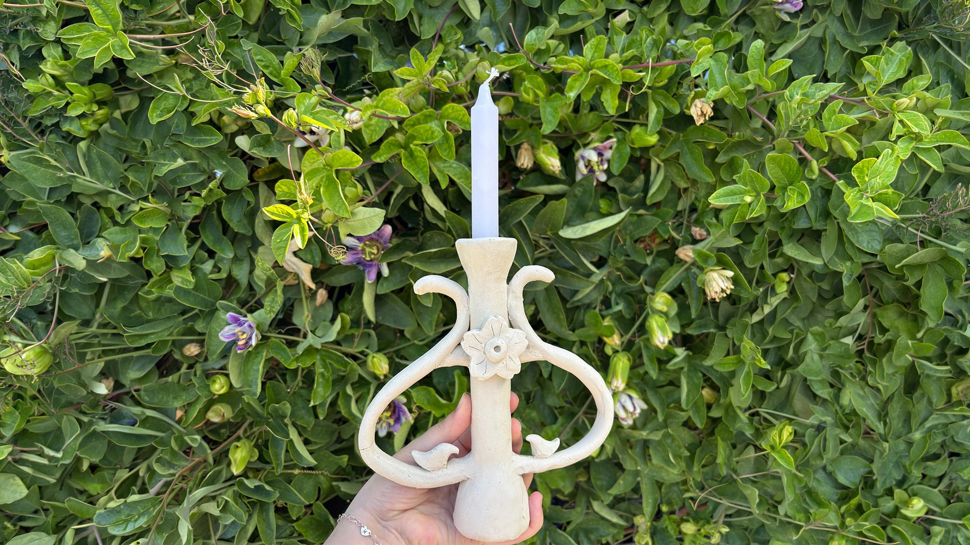 Hand holds floral ceramic candleholder before flowering vines.