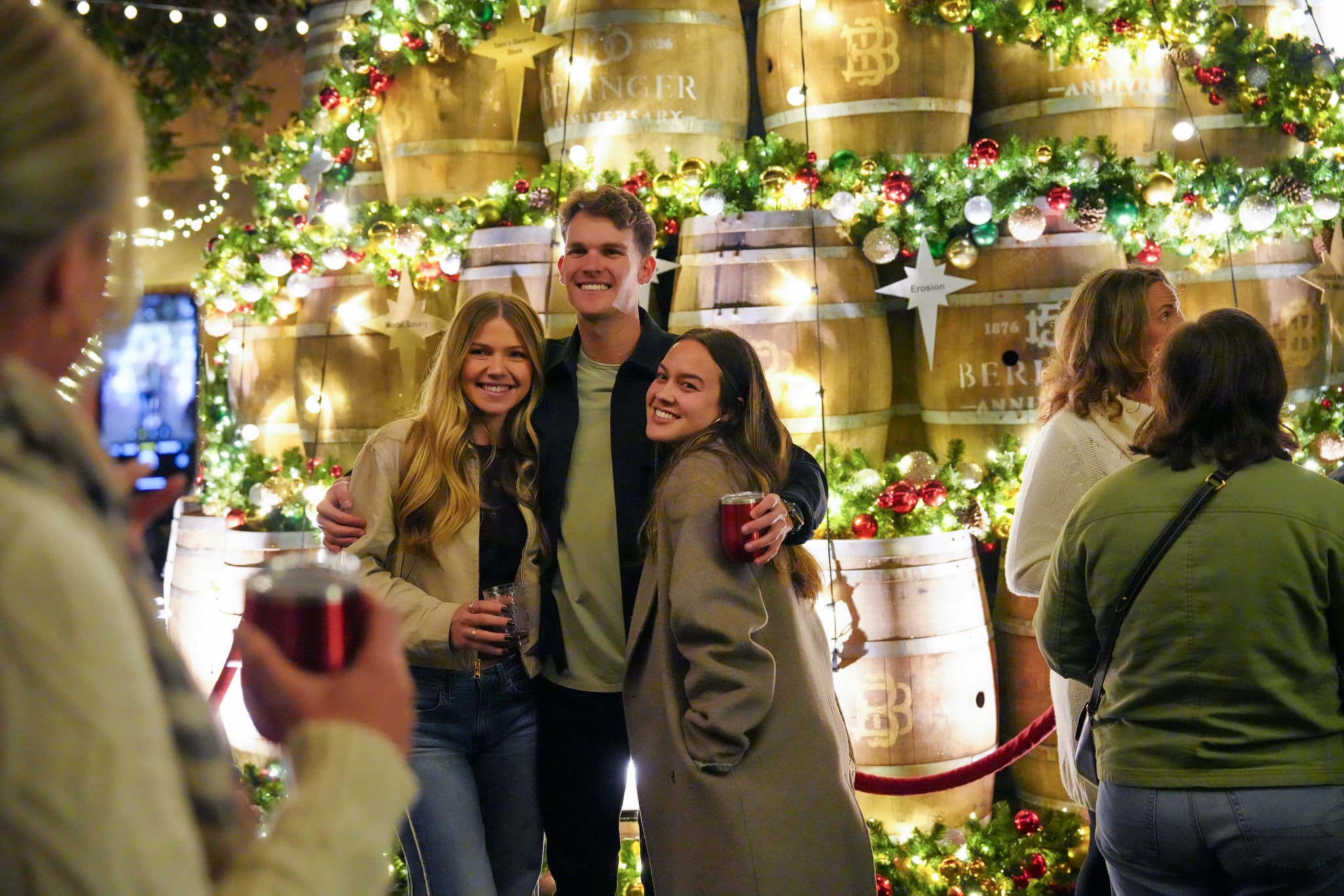Friends pose before festive, illuminated whiskey barrel display.