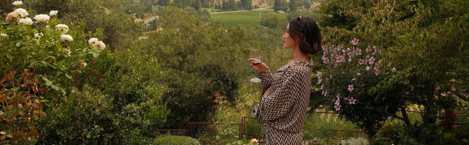 Woman holding wine overlooking lush vineyard garden