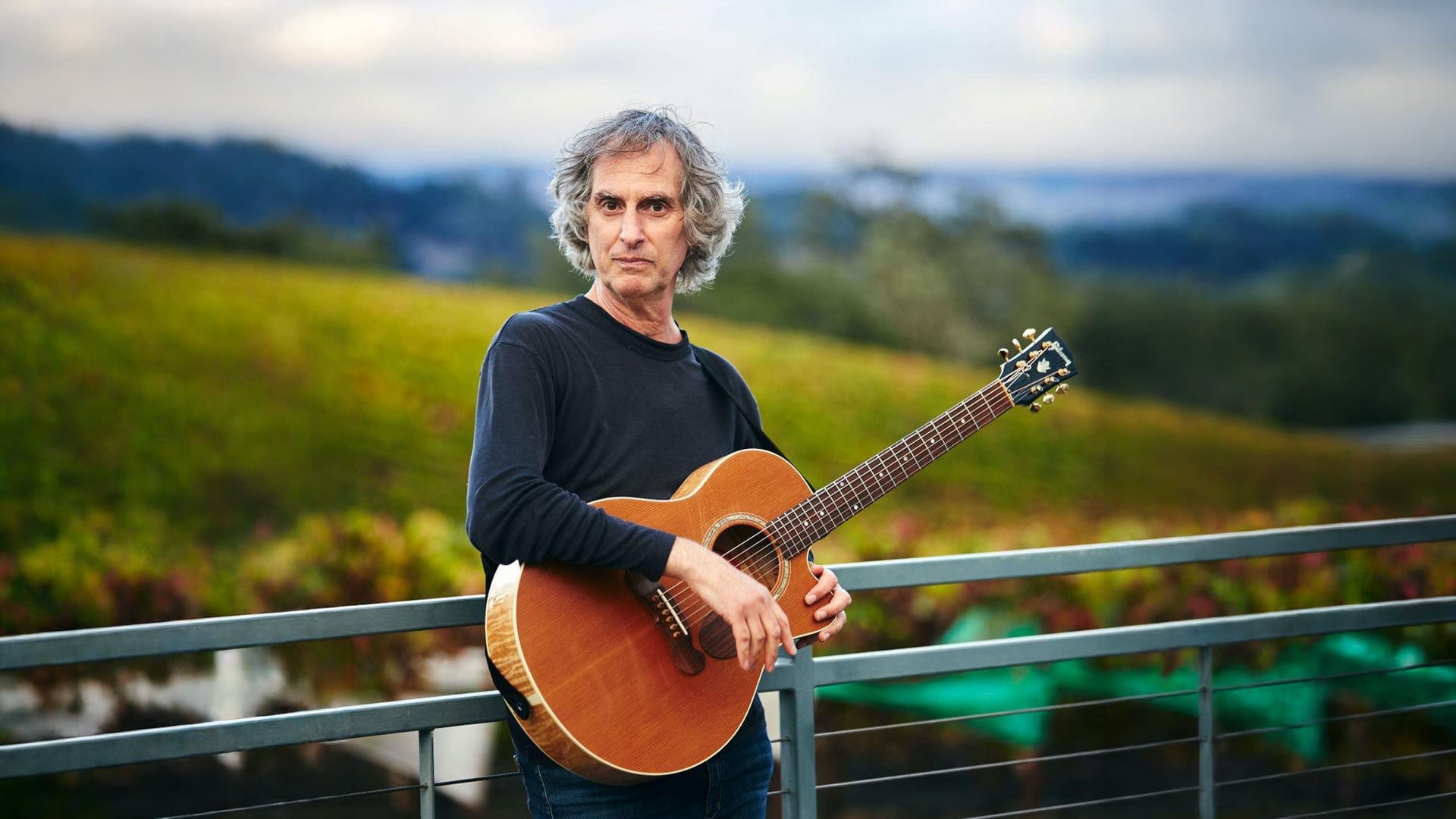 Gray-haired man holding acoustic guitar on outdoor balcony