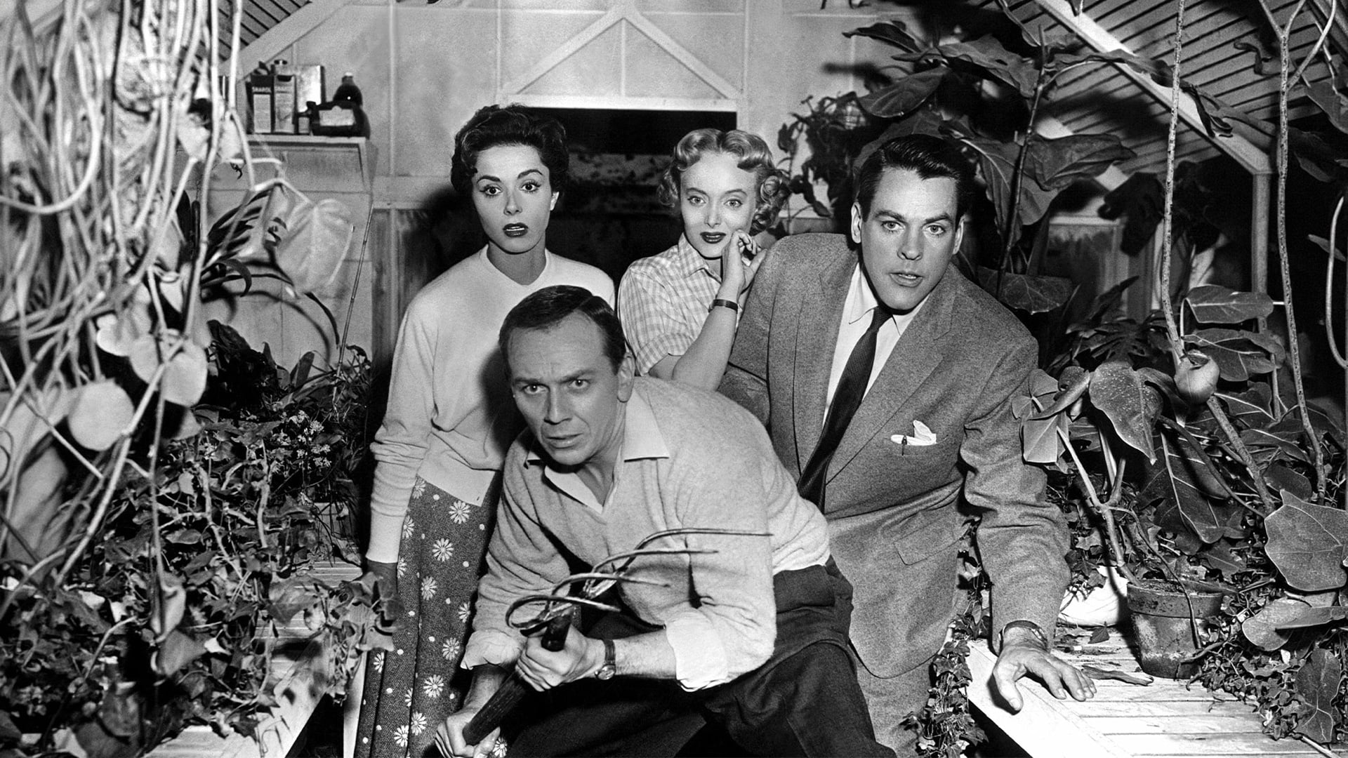 Four startled people crouch among dense indoor plants.