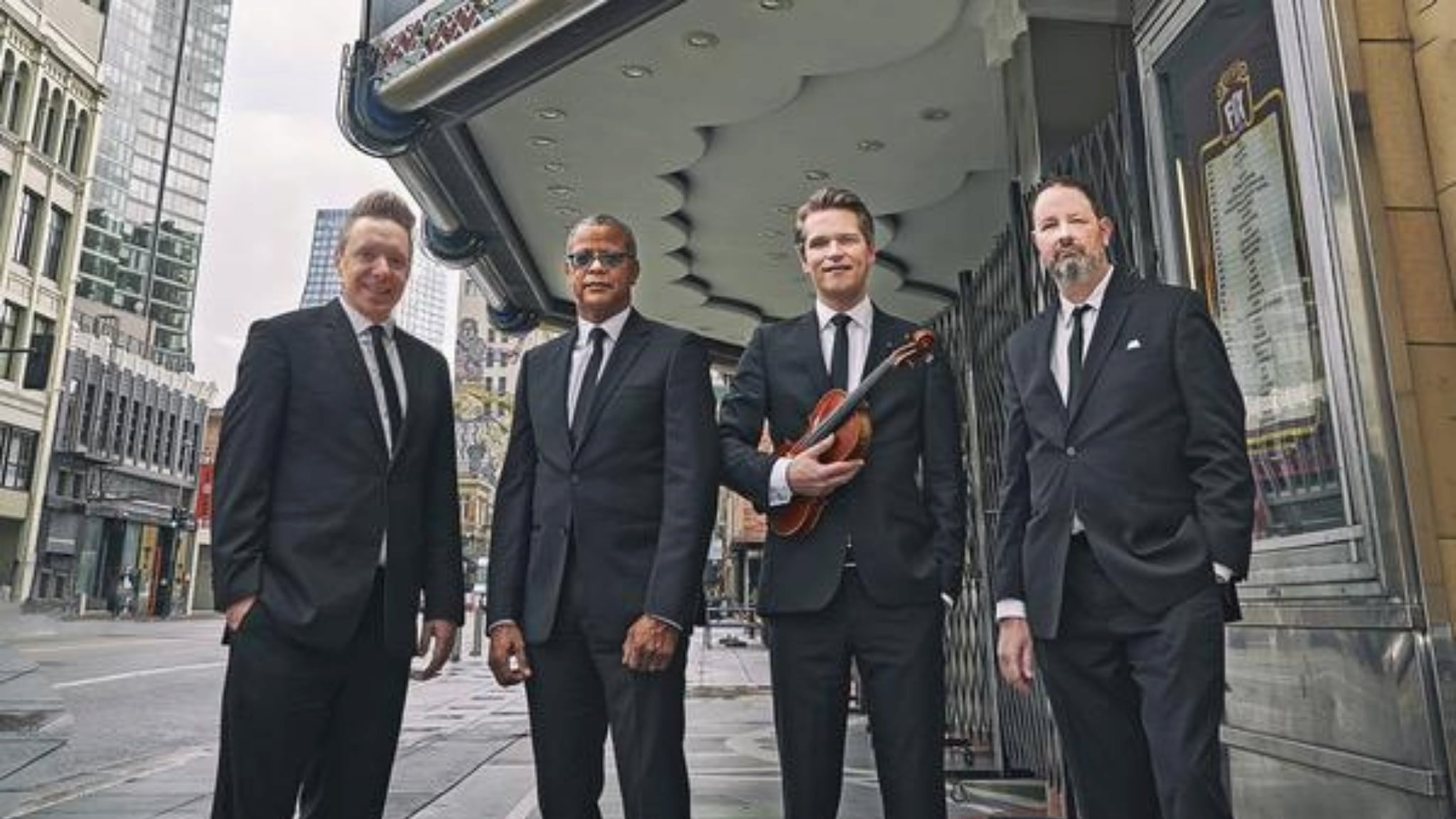 Four men in suits outside theater, one holding violin